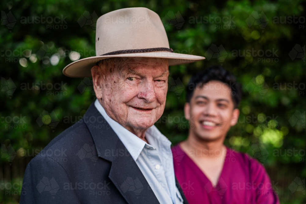 Portrait of an elderly man in a hat smiling and standing outdoors with male healthcare worker - Australian Stock Image