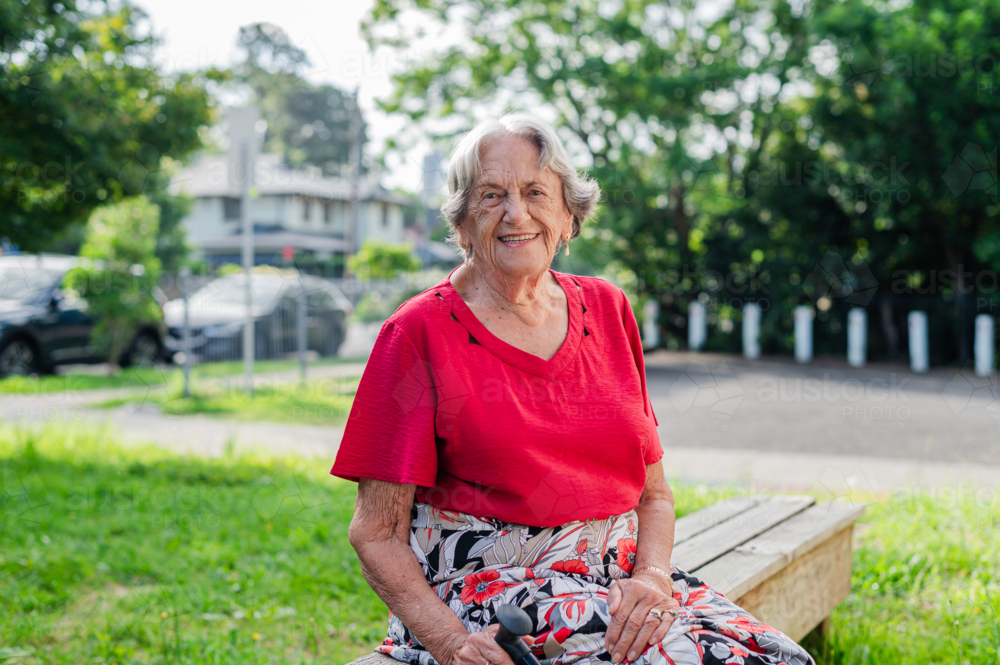 Portrait of an elderly woman in red blouse sitting on wooden bench outdoors smiling - Australian Stock Image