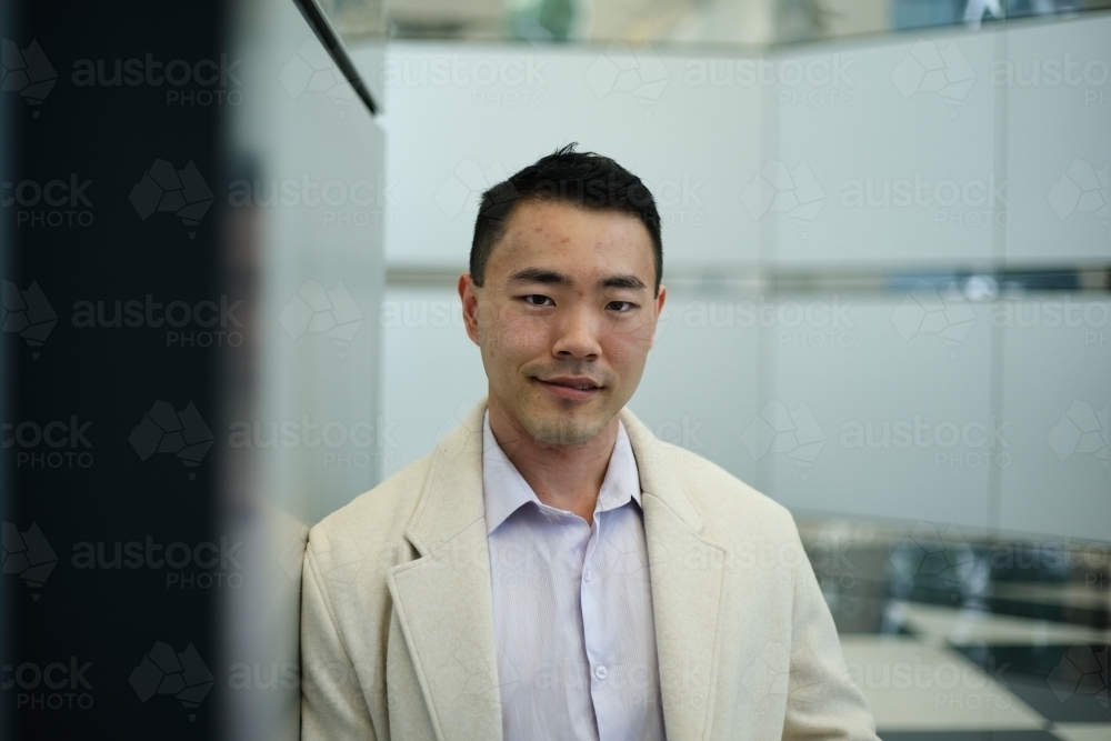 Portrait of Asian man in modern workplace leaning against wall - Australian Stock Image