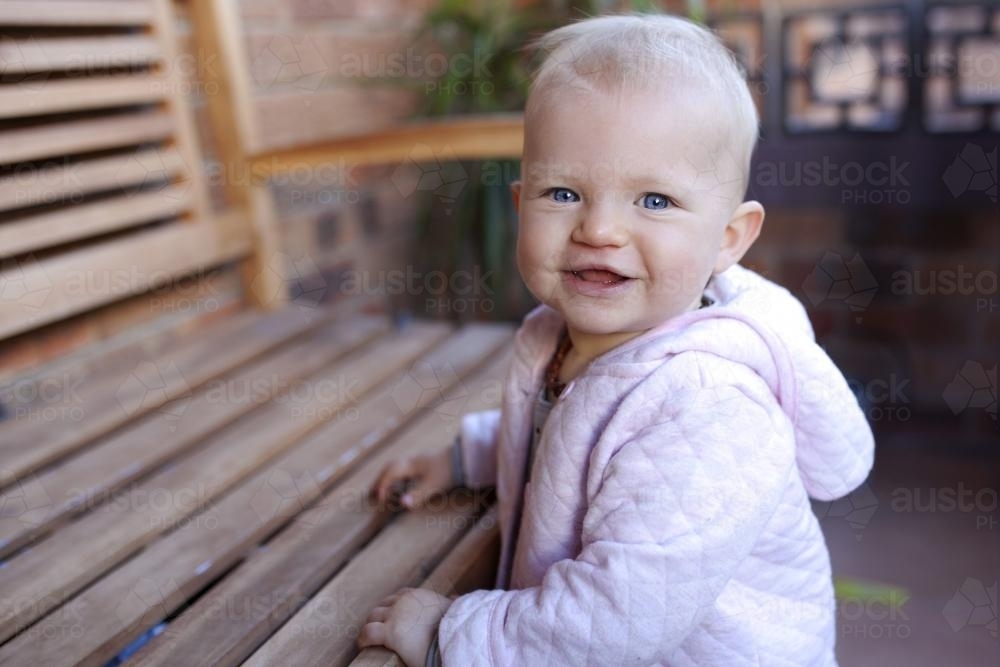 Portrait of baby girl smiling, standing at a wooden seat - Australian Stock Image