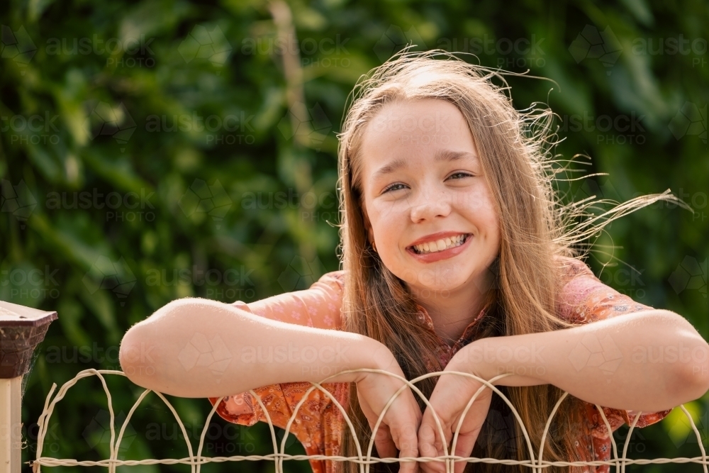 Portrait of cheerful pre-teen girl leaning on wire fence in country town - Australian Stock Image