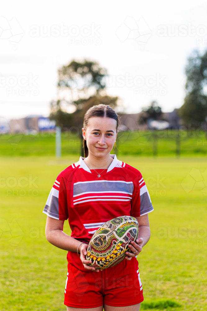 Portrait of female Aboriginal teenager holding AFL footy ball on sports ground field - Australian Stock Image
