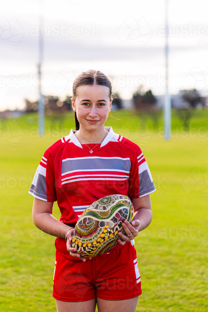 Portrait of female Aboriginal teenager holding AFL footy ball on sports ground field - Australian Stock Image