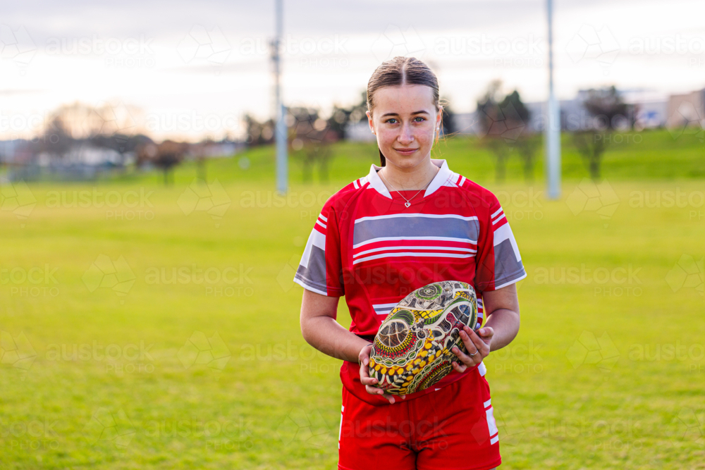 Portrait of female Aboriginal teenager holding AFL footy ball on sports ground field - Australian Stock Image