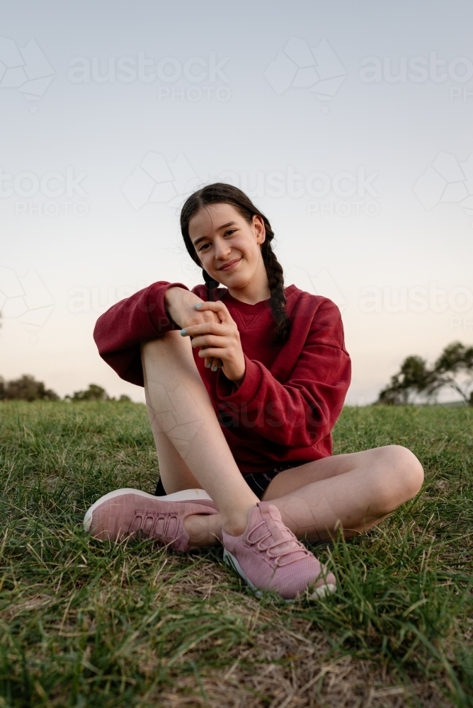 Portrait of happy young adolescent girl with braided hair sitting cross legged on grass - Australian Stock Image