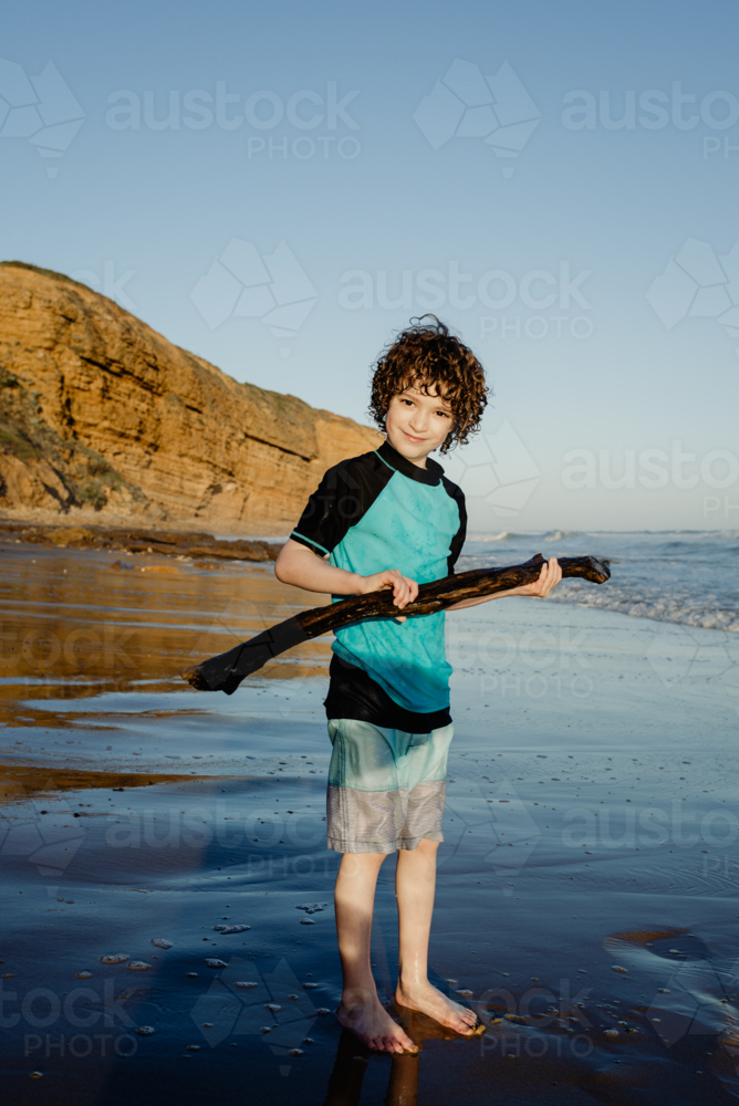 Portrait of happy young boy with curly hair holding a stick on a beach - Australian Stock Image