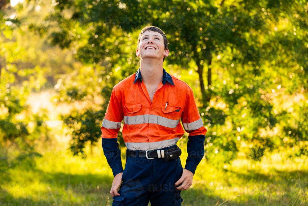 Portrait of happy young tradesman apprentice in high vis workwear looking upwards at sky - Australian Stock Image