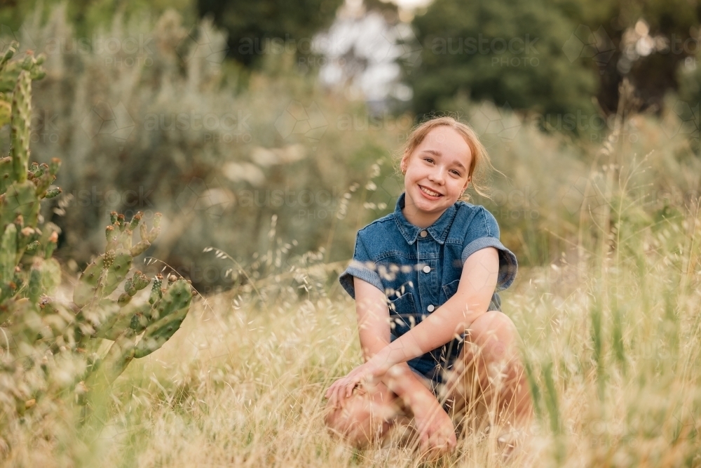 Portrait of pre-teen girl sitting in long grass in natural outdoor setting - Australian Stock Image