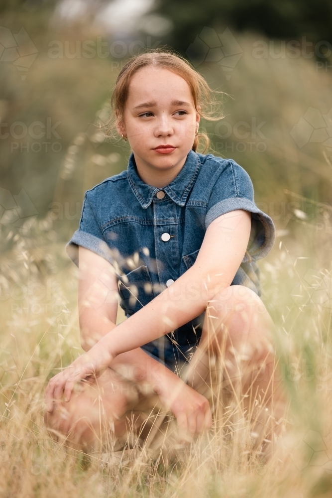 Portrait of pre-teen girl sitting in long grass in natural outdoor setting - Australian Stock Image