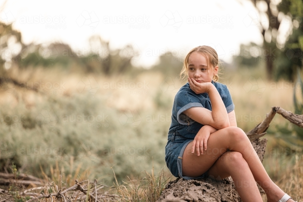Portrait of pre-teen girl sitting on log in natural setting with thoughtful expression - Australian Stock Image