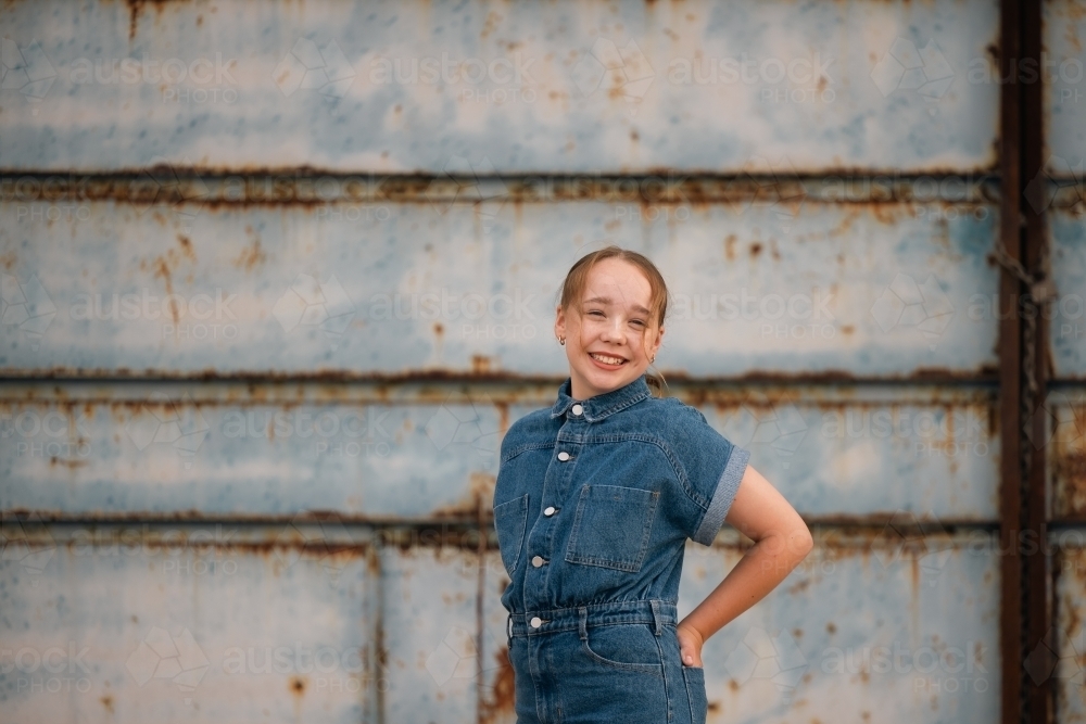 Portrait of pre-teen girl wearing denim jump suit in front of large blue barn door - Australian Stock Image