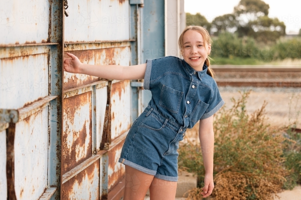 Portrait of pre-teen girl wearing denim jump suit in front of large blue barn door - Australian Stock Image
