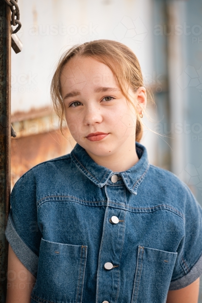 Portrait of pre-teen girl wearing denim jump suit in front of large blue barn door - Australian Stock Image