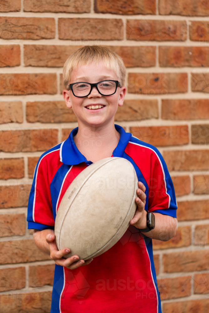 Portrait of primary school boy holding footy ball by brick wall - Australian Stock Image