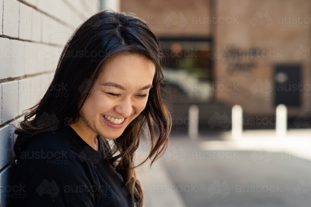 portrait of smiling asian woman, casual portrait leaning against a brick wall - Australian Stock Image