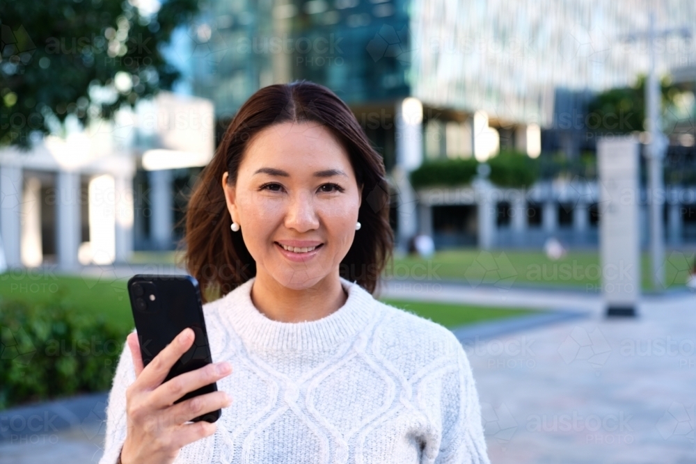 Portrait of smiling woman holding phone next to park - Australian Stock Image
