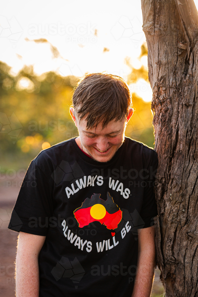 Portrait of teenaged aboriginal boy smiling by tree in bushland at sunset - Australian Stock Image