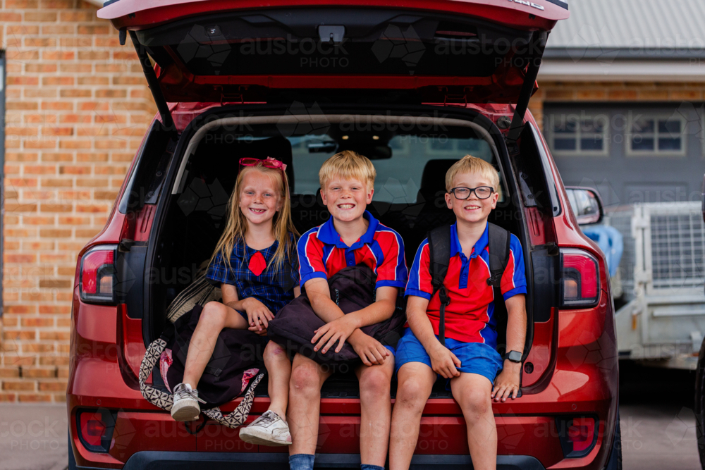 portrait of three siblings sitting together in car boot ready for school drop off smiling - Australian Stock Image