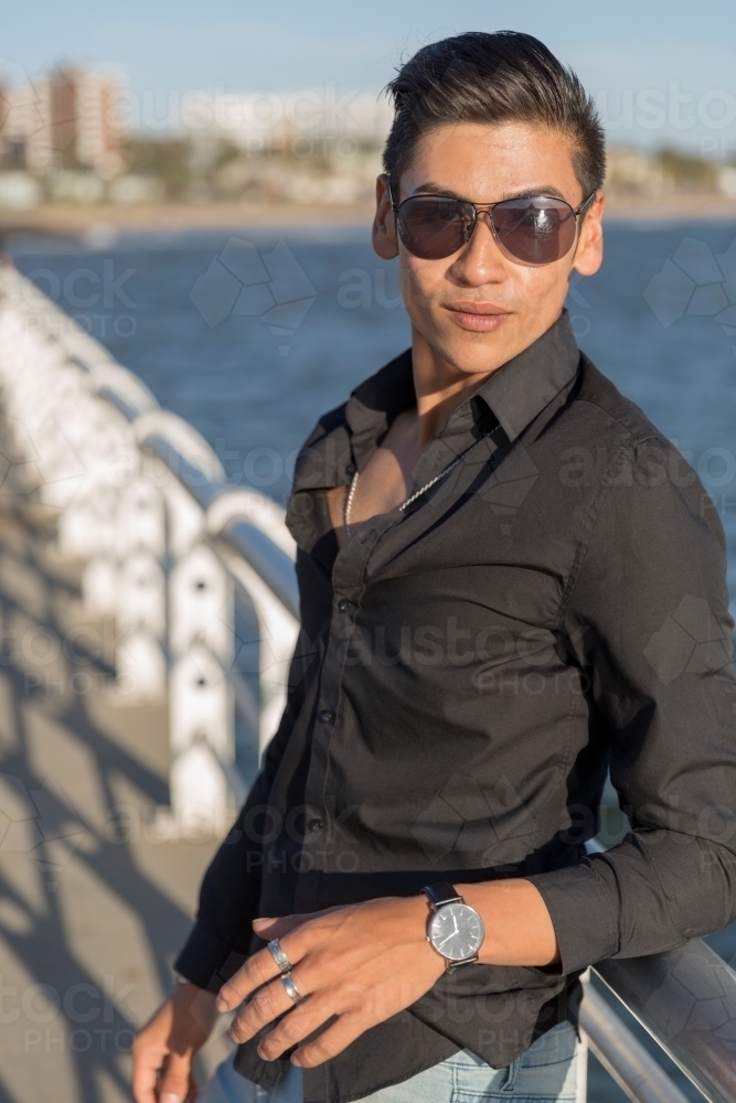 Portrait of trendy young man leaning on railing of pier with sunglasses - Australian Stock Image
