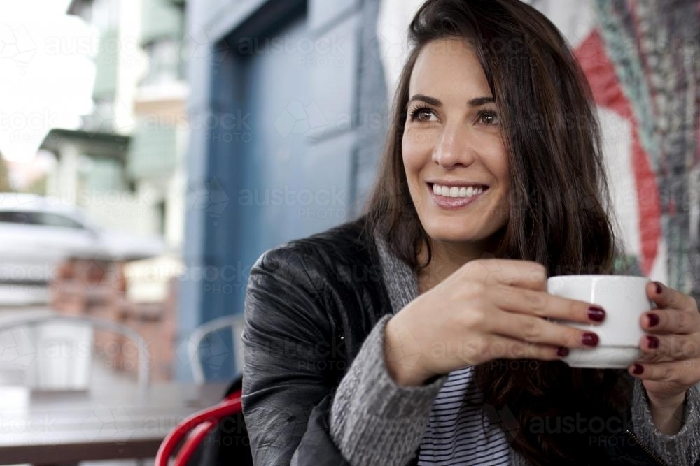 Portrait of woman sitting at a cafe smiling and holding a cup - Australian Stock Image
