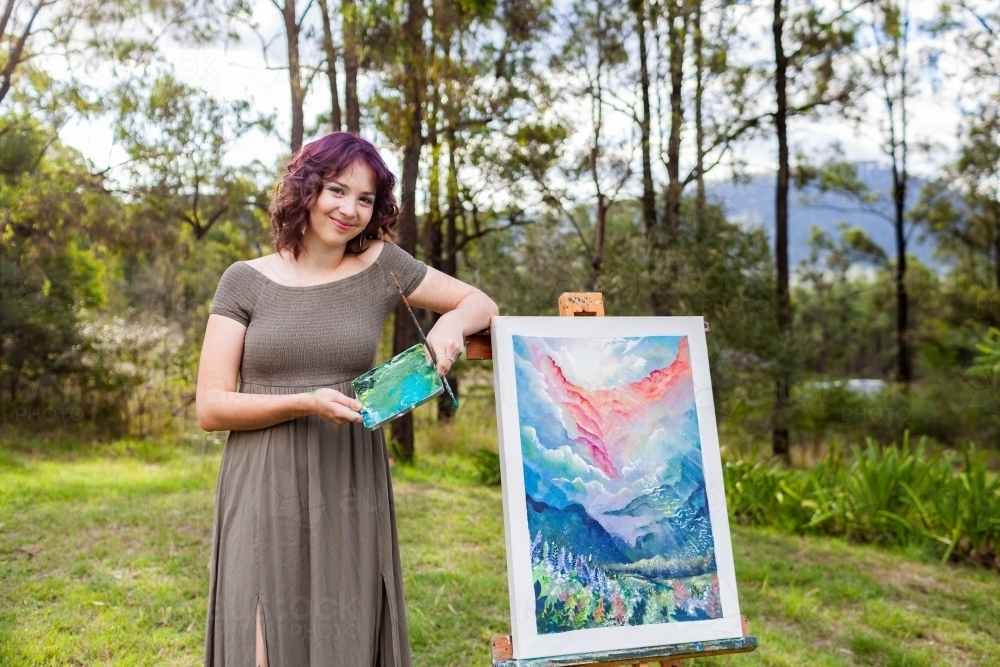 Portrait of young artist standing beside artwork of dreamy land and cloudscape painting - Australian Stock Image