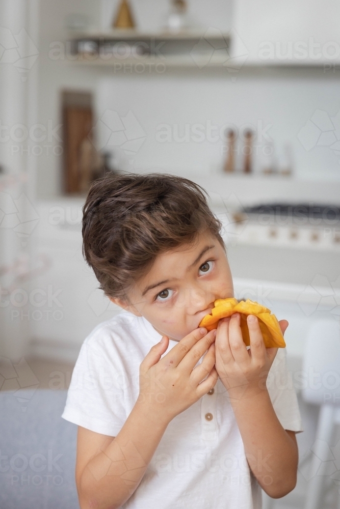 Portrait of young boy eating slice of mango in kitchen - Australian Stock Image