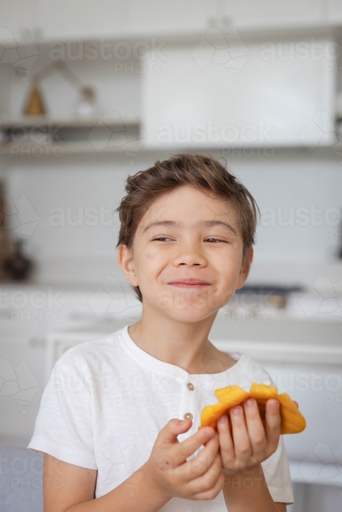 Portrait of young boy eating slice of mango in kitchen - Australian Stock Image