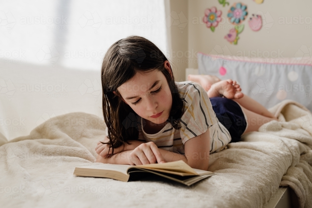 Portrait of young preteen girl in her bedroom lying on her bed reading a book by a sunny window - Australian Stock Image