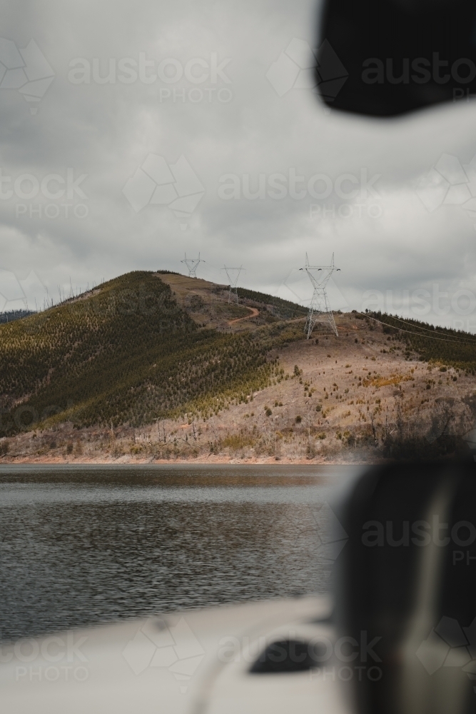 Powerlines on a mountain through a pine forest clearing with a lake in the foreground - Australian Stock Image