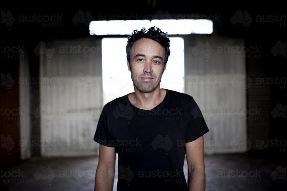 Pportrait of a man wearing a black t-shirt in a dimly lit shed - Australian Stock Image
