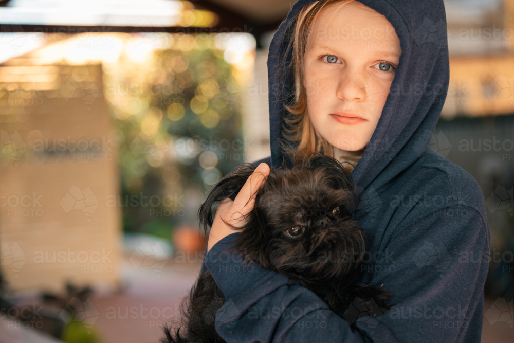 Pre-teen boy wearing hoodie snuggling little dog - Australian Stock Image