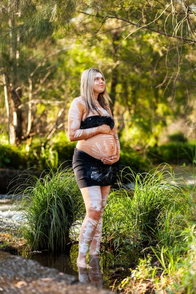Pregnant Aboriginal lady in ochre body paint standing by water in bushland - Australian Stock Image
