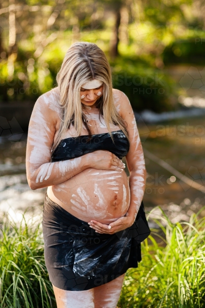 Pregnant Aboriginal lady in ochre body paint standing by water in bushland - Australian Stock Image
