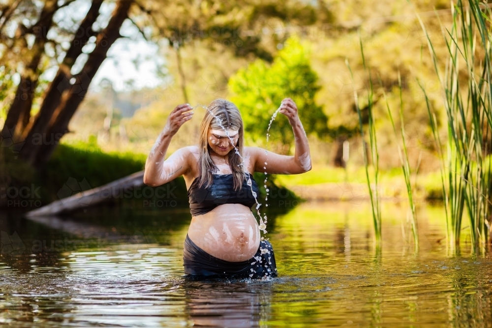 Pregnant Aboriginal woman in traditional body paint splashing streams of water droplets in creek - Australian Stock Image