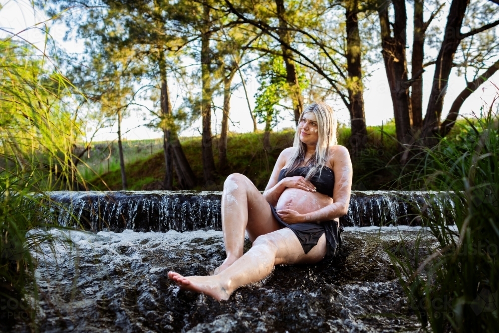 Pregnant aboriginal woman sitting in flowing water bonding with her unborn child - Australian Stock Image