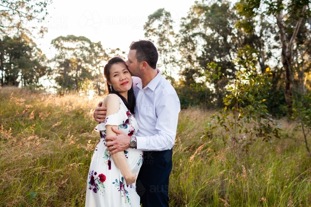 Pregnant Asian mum to be with loving husband kissing her in aussie bushland - Australian Stock Image