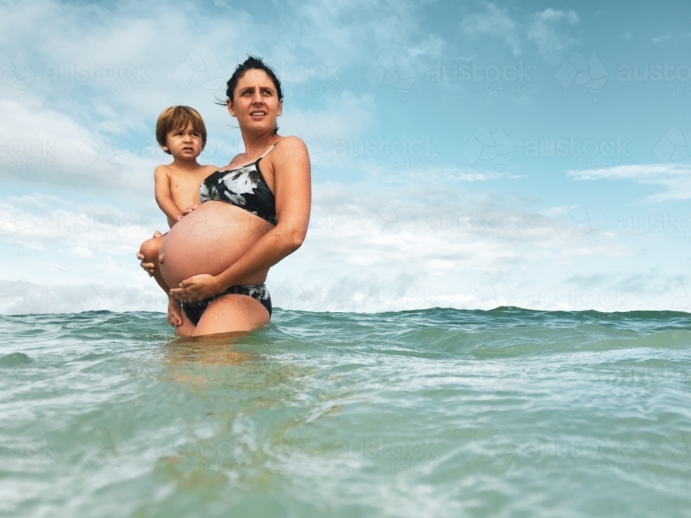 Pregnant woman holding toddler, standing in the ocean - Australian Stock Image