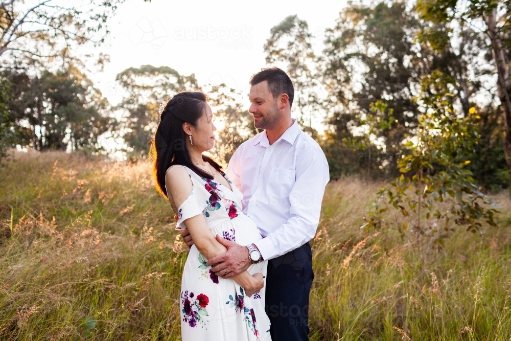 Pregnant woman of Asian ethnicity with happy husband standing together outside in australian light - Australian Stock Image