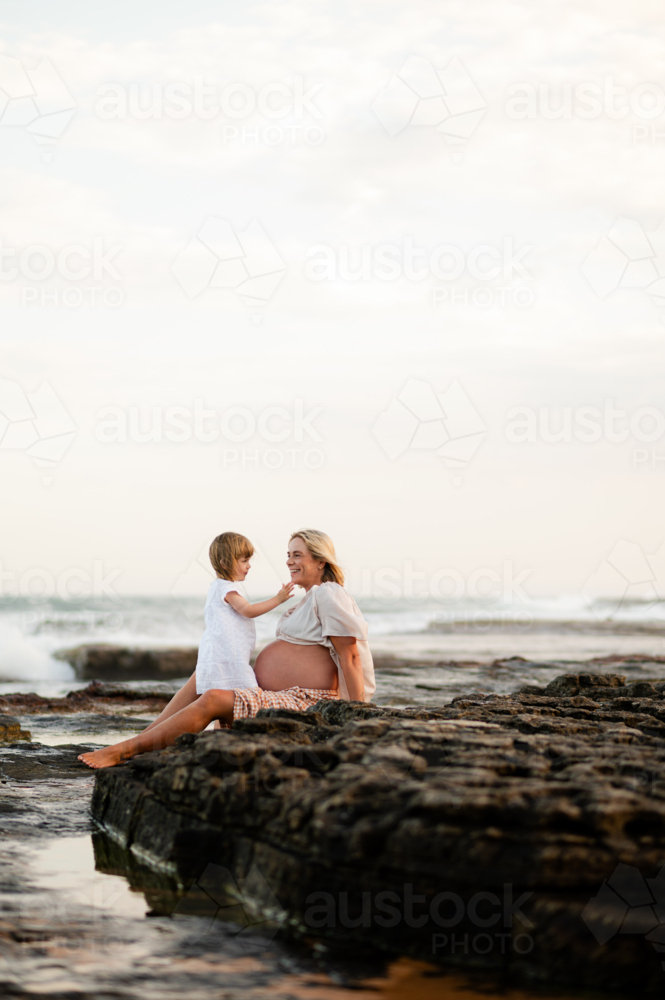Pregnant woman seated on rocks at sunset with child playing at ocean's edge - Australian Stock Image