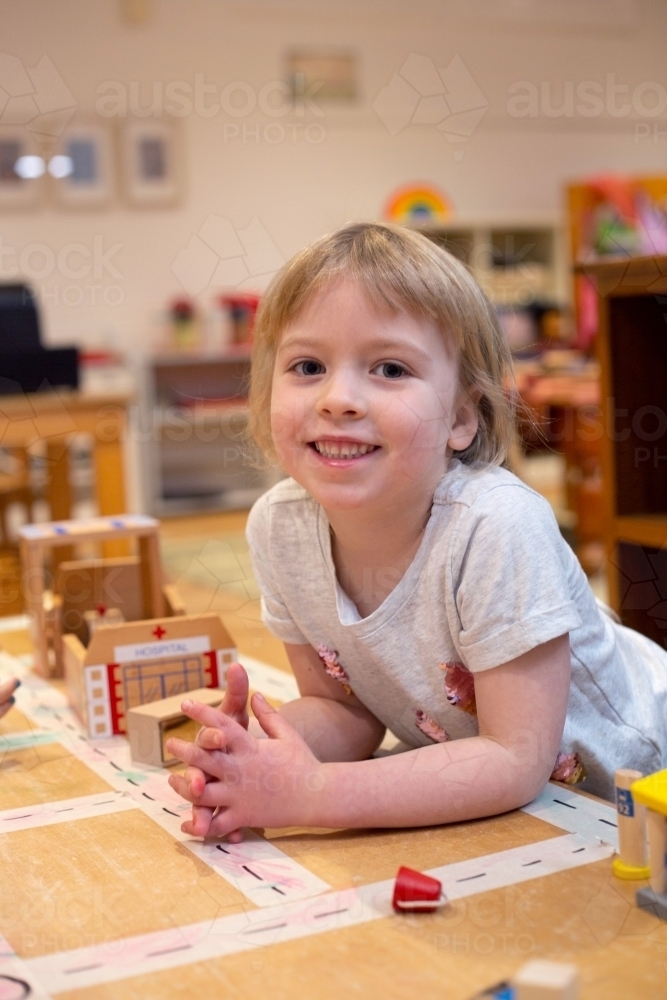 Preschool girl leaning on a desk and smiling in play room - Australian Stock Image