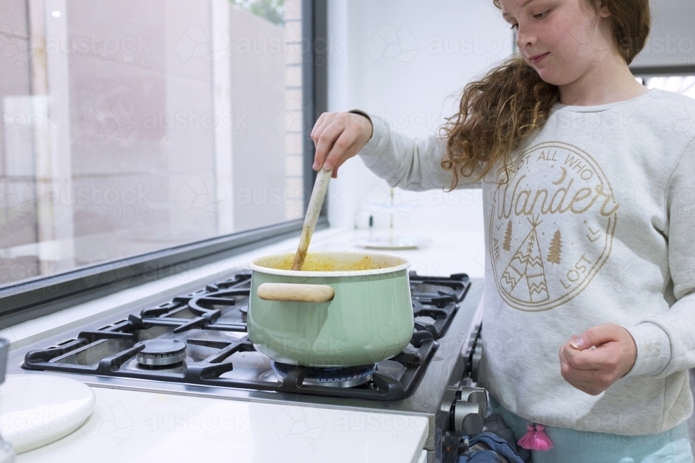 Preteen girl helping to cook over a saucepan on a stove in the kitchen - Australian Stock Image