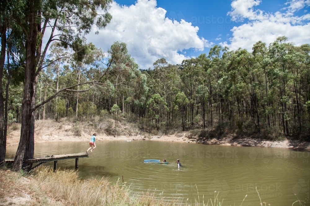 Preteen girl jumping off jetty into rural dam to swim with friends - Australian Stock Image