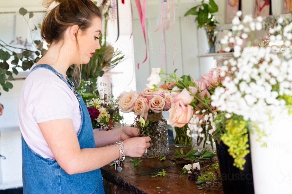 Pretty young florist preparing wedding buttonholes at a work bench full of flowers - Australian Stock Image