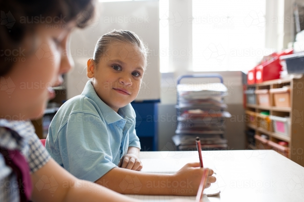 Primary school girl facing the camera in a classroom whilst writing - Australian Stock Image