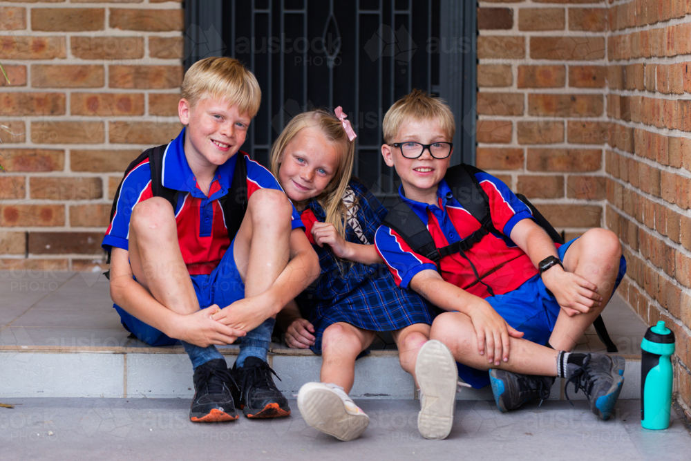 Primary school students sitting together on front doorstep of home ready to go to school - Australian Stock Image