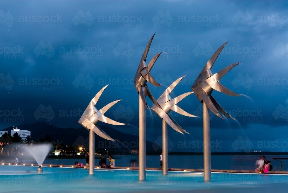 Public pool with fish statues, people in background, in night light - Australian Stock Image