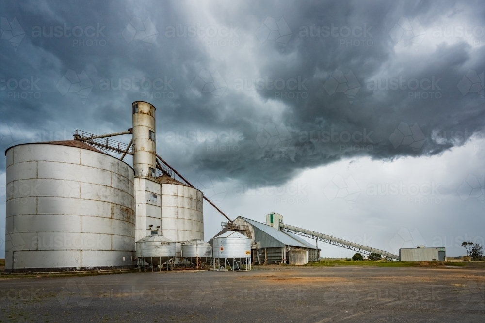 Rain falling from a dramatic stormfront over grey grain storage silos in a rural setting - Australian Stock Image