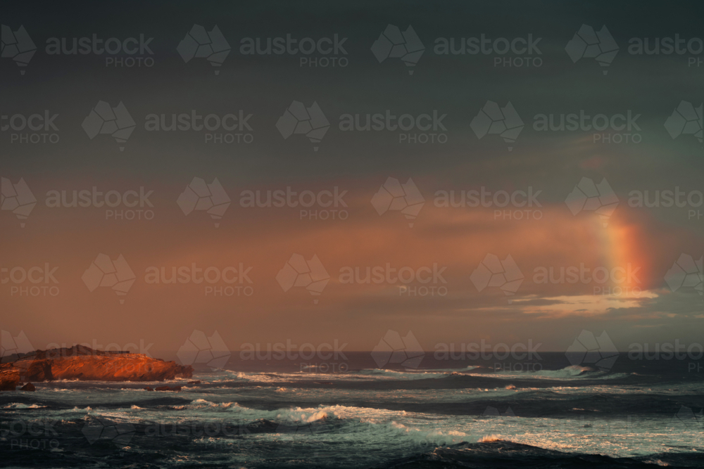Rainbow above cliffs with waves at glowing sunset - Australian Stock Image