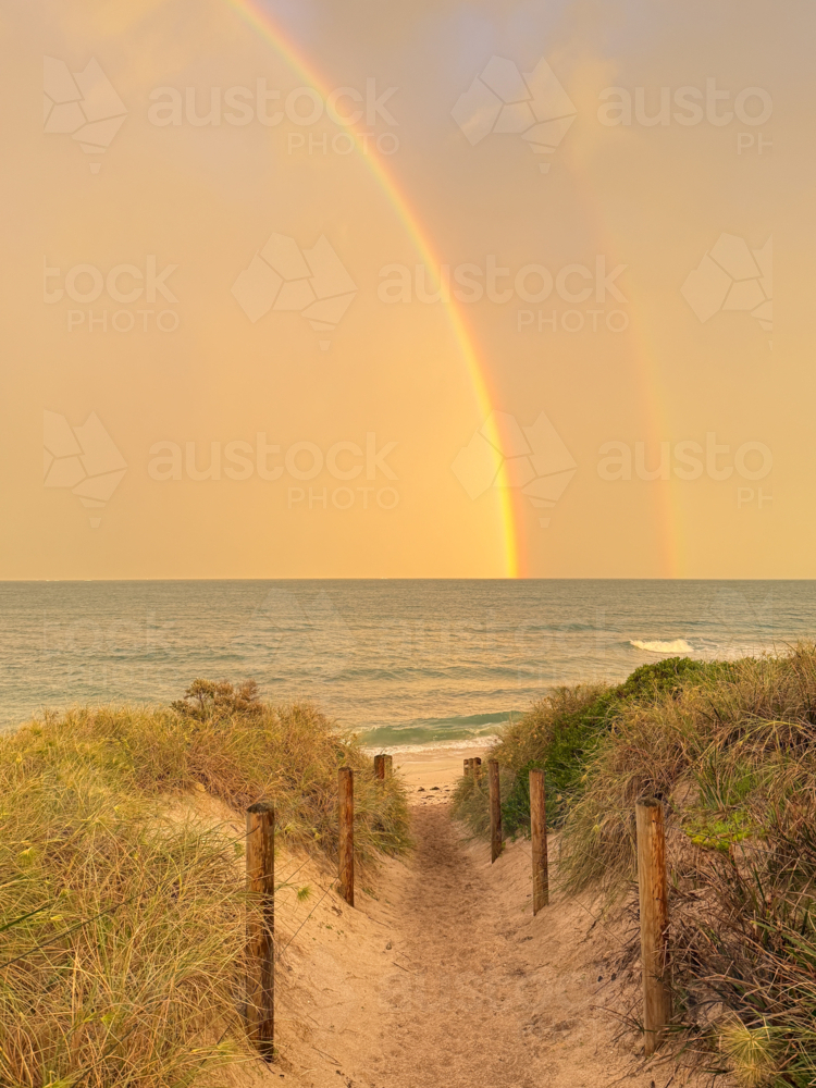 Rainbow sitting over ocean seen through fenced beach walkway - Australian Stock Image