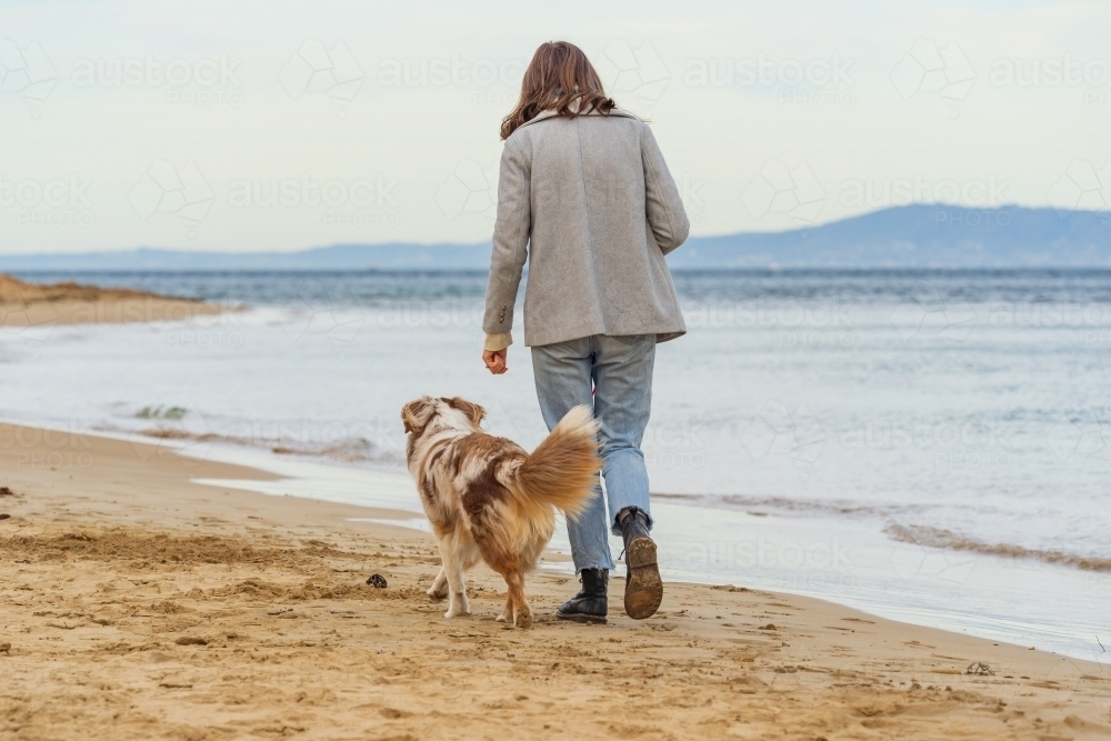 Rear view of a young woman walking a fluffy dog on a beach on an overcast day - Australian Stock Image